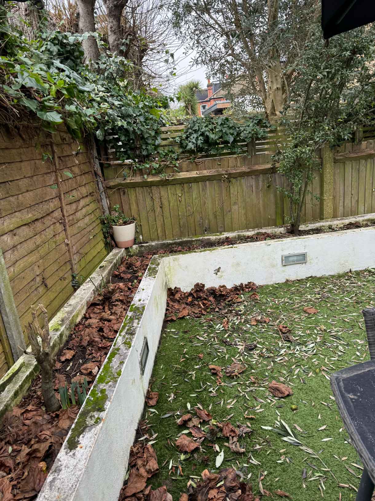 Garden before renovation with old fencing, patchy grass, and white concrete planters.