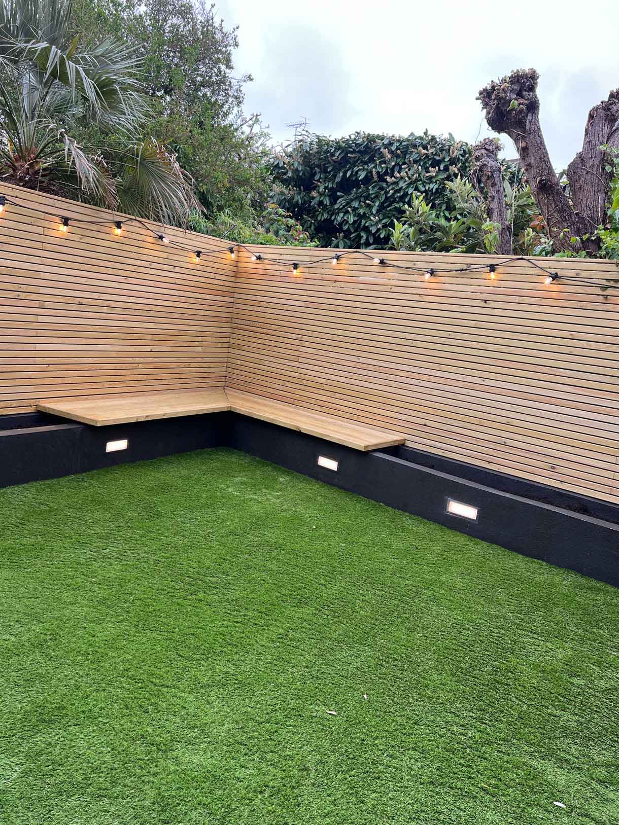 Corner of courtyard garden with seating, lawn, and decorative grasses in planters.