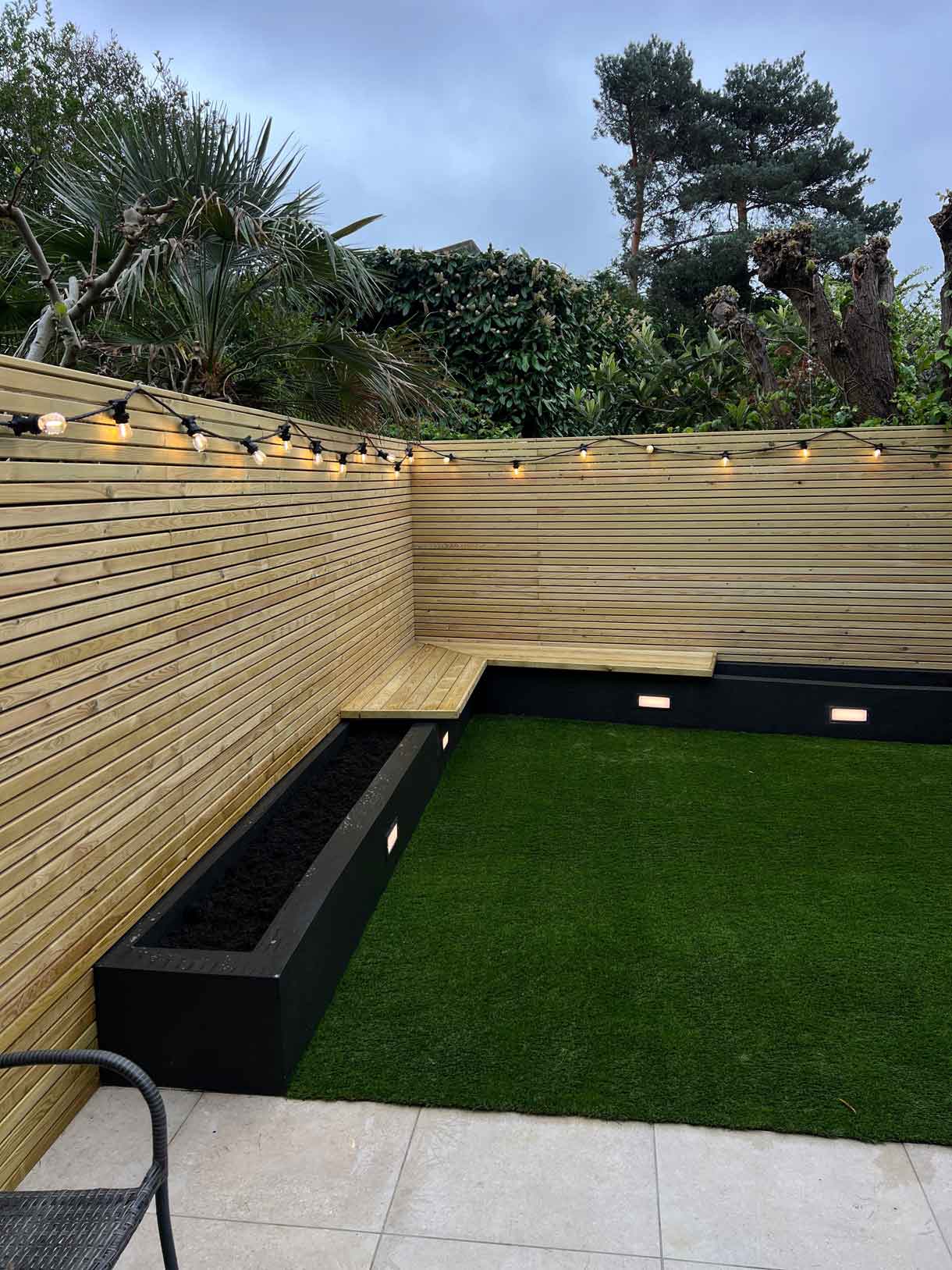 Evening view of courtyard garden with string lights, lawn, and timber seating.