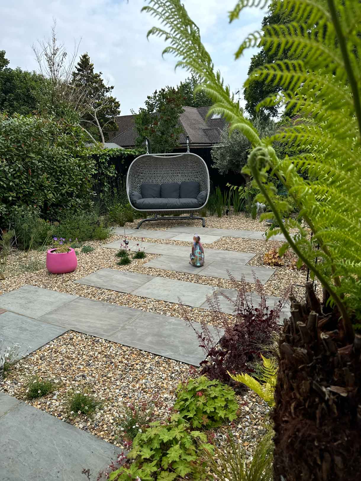 Gravel garden with staggered stone paving, colourful plants, and a grey outdoor swing seat, with a pink planter adding contrast in the corner.