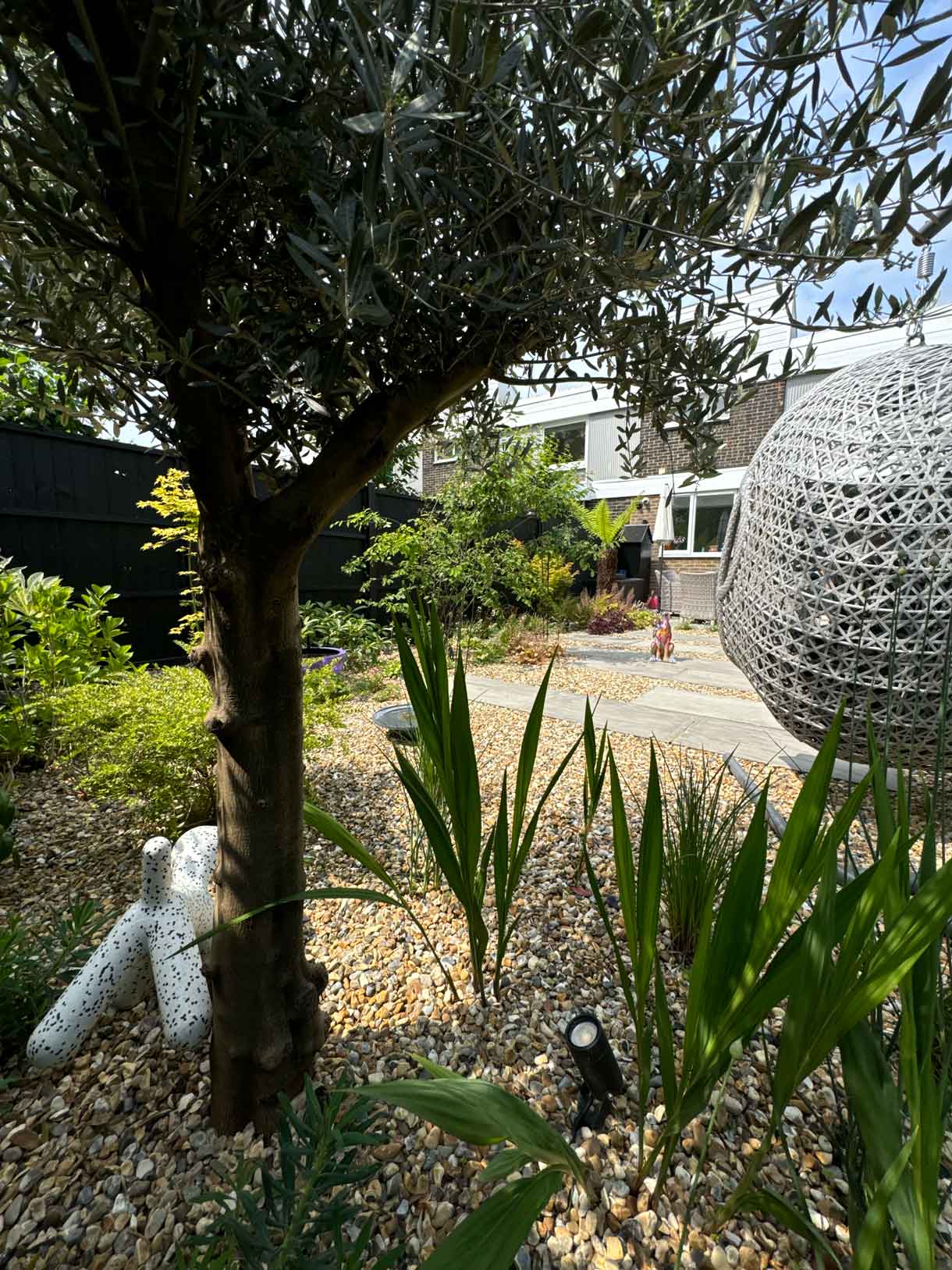 View of gravel garden through tree planting, with a grey swing seat visible in the background.