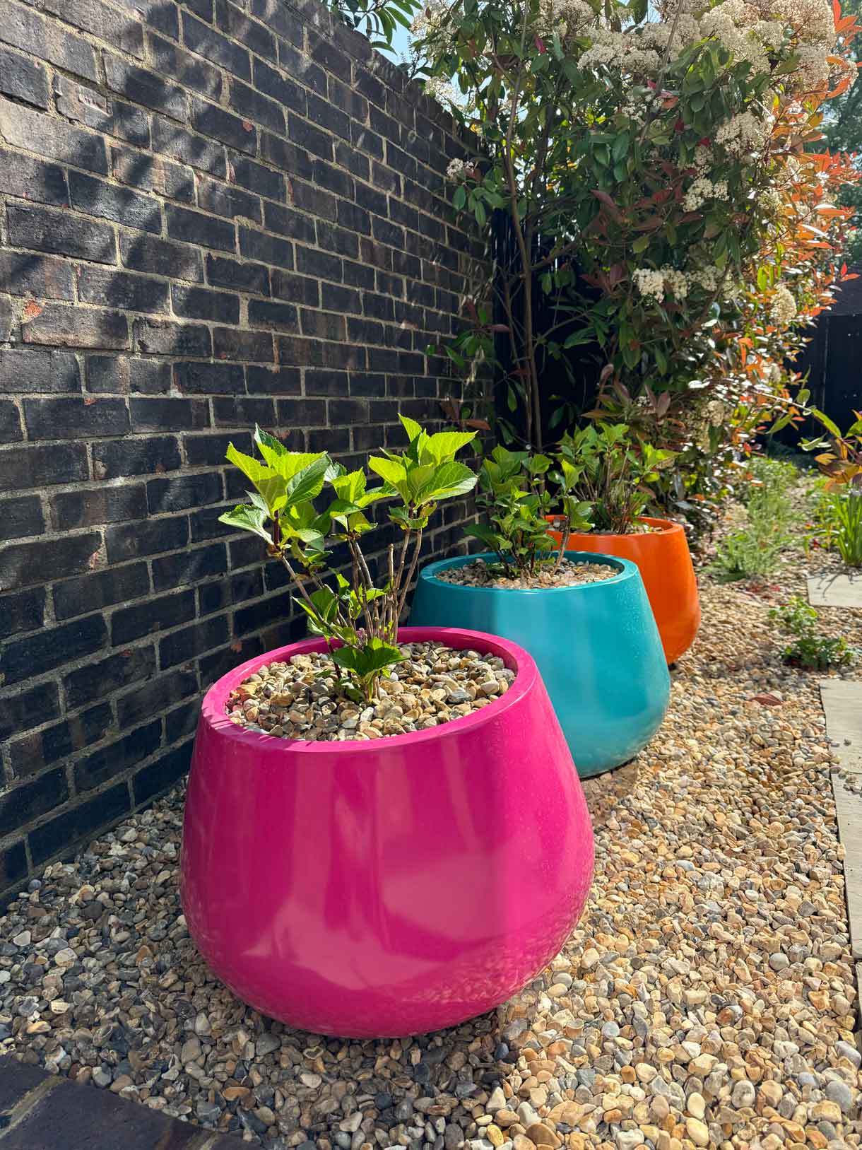 Row of colourful planters in pink, turquoise, and orange set against a brick wall with gravel and planting
