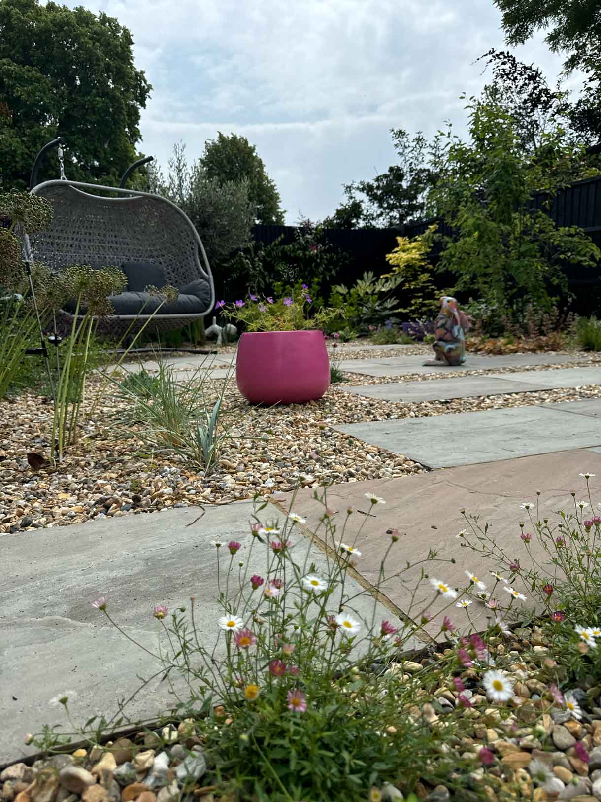 Gravel garden with staggered paving and a bright pink planter, featuring a swing seat in the background
