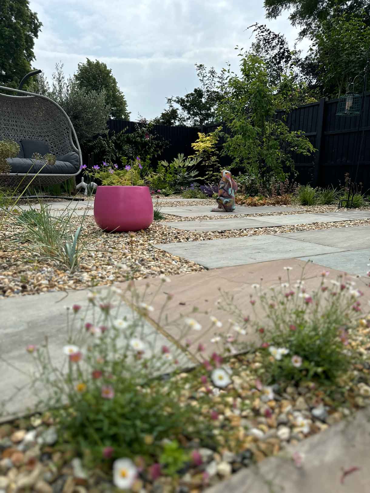 Pink planter and staggered paving in gravel garden with swing seat in the background.