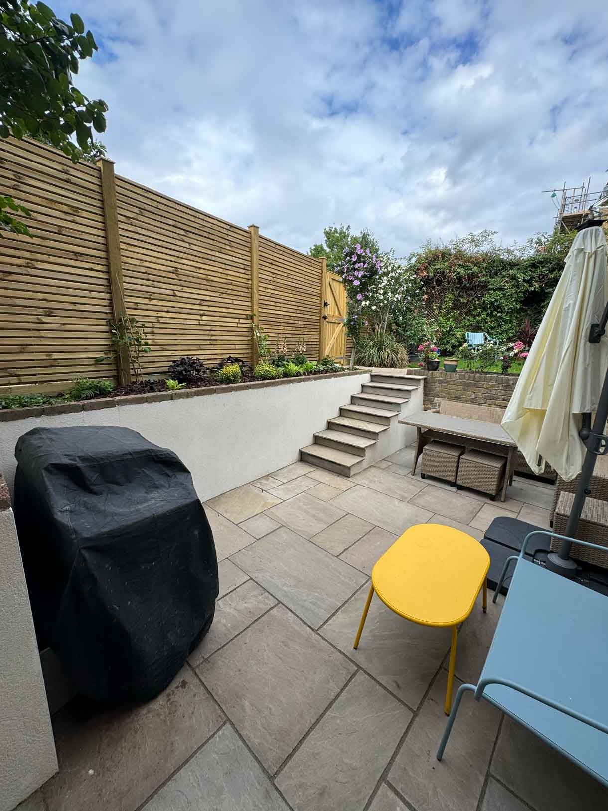Paved basement patio with barbecue cover, yellow stool and steps leading up to a fenced garden.